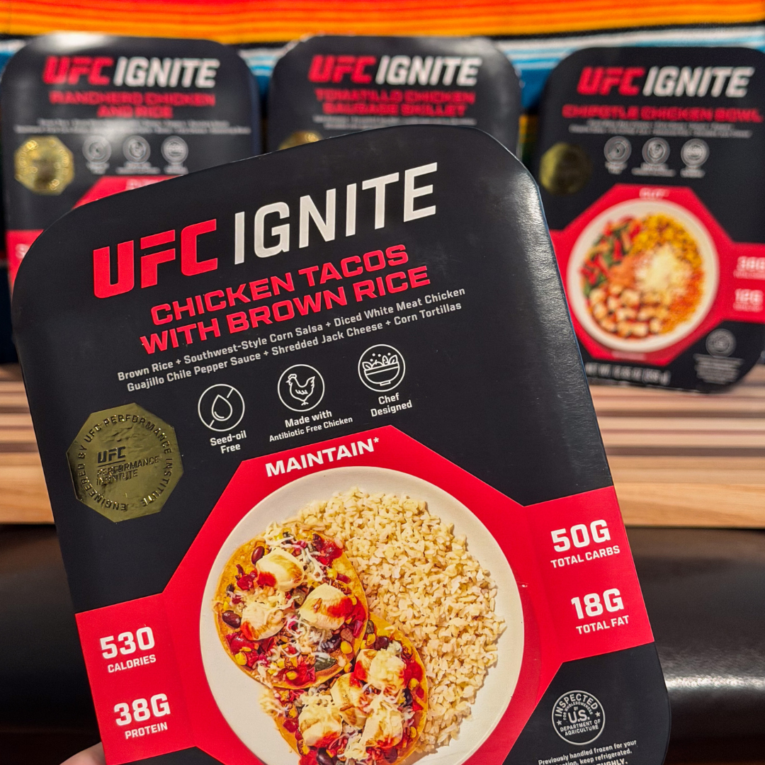 Hand holding the UFC Ignite Chicken Tacos with Brown Rice meal with the other three Noche UFC Box meals behind